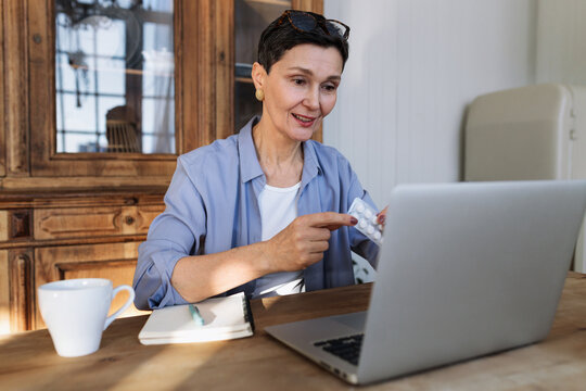 Senior intelligent female GP giving medical consultations in real time using laptop sitting in her cabinet against antique wardrobe, holding pill pack and giving instructions of its intake