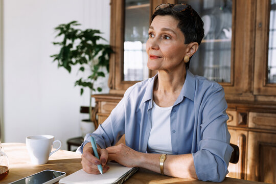 Senior lady in casual clothes writing in thought or mood diary to reveal her true emotions and feelings, writing gratitude journal as self-care or cognitive behavioral tool sitting in living-room