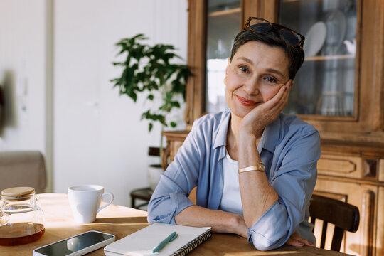 Happy elderly woman enjoying her morning sitting at table with cup of coffee and copybook with pen, going to make to do list for day or week plan, smiling at camera with her chin on hand