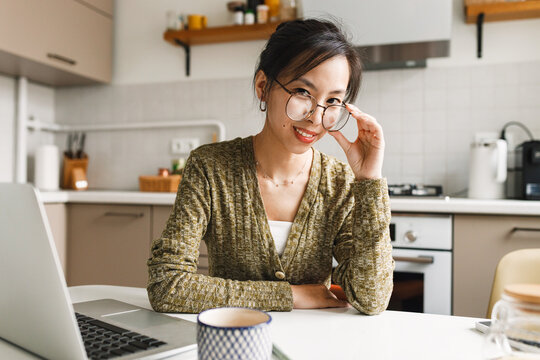 Young asian teacher in stylish glasses sitting at kitchen table, having break while preparing for online lesson, reviewing materials on laptop. Concept of remote work in education and tutoring
