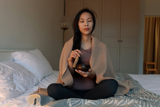 Asian woman meditating with singing bowl on bed at morning with blanket on shoulders, eyes closed, feeling calm and inner peace through sound vibration. Mindfulness practice for energy and wellness