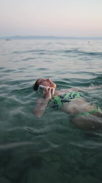 Vertical screen woman floats on her back in calm sea while talking on smartphone showing phone addiction and inability to disconnect from calls even during relaxing summer swim