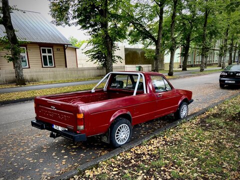 Rear three-quarter view of a classic red Volkswagen Caddy Mk1 pickup with roll bar, parked on an autumn street in Estonia surrounded by fallen yellow leaves and wooden houses.