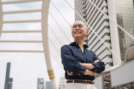 Confident bald business woman in glasses smiles looking up, arms crossed, wearing navy blue shirt and white trousers on modern city skywalk with skyscrapers, urban lifestyle portrait