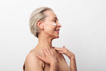 Fototapeta premium Smiling mature woman with short hair applying moisturizer on face and shoulders isolated on white. Concept of skincare routine, natural beauty, anti aging, wellness, and self care.
