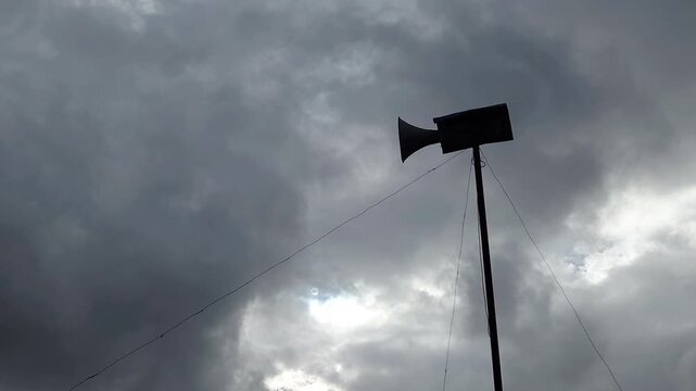 Low angle view of a loudspeaker horn mounted on a tall pole against a dramatic overcast sky with dark clouds moving in the wind, symbolizing a call to prayer, public announcement, or warning.