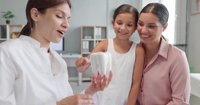 Dentist guides child and mother on toothbrush hygiene. In a bright clinic, a pediatric dentist explains brushing in consultation using a tooth model. Strong family teeth prevention concept.