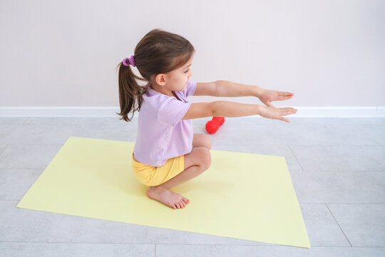 Young Girl Squats On Yoga Mat With Arms Stretched Forward During Home Exercise Practice, Showing Balance, Flexibility, And Focus In Bright Minimal Room With Massage Ball And Soft Natural