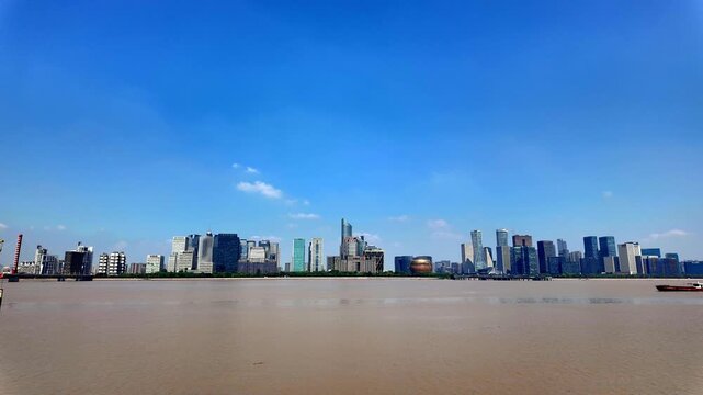 A panoramic view of a modern urban skyline along a wide, calm river under a bright clear blue sky on a sunny day.