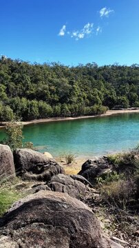 Vertical shot of sunlight warms the large granite boulders along the edge of a quiet lake nestled within a lush green forest on a bright afternoon