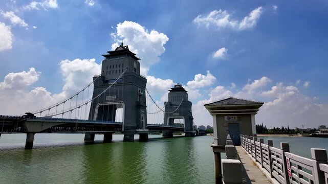 A suspension bridge with traditional styled towers spans a green river under a bright blue sky with white clouds.