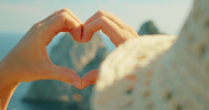 Hands forming heart shape framing distant Faraglioni sea cliffs over calm blue Mediterranean waters in Capri