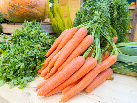 Fresh bundle of young carrots with leafy tops arranged on wooden table alongside parsley and zucchini in bright outdoor market setting, with carrot, leafy, parsley, zucchini detail