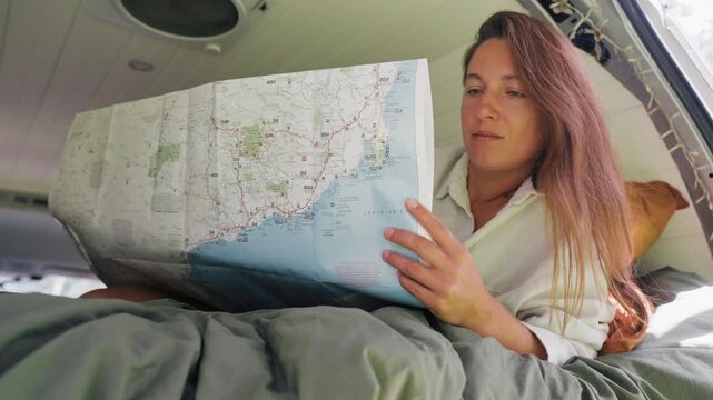 Woman resting on a bed inside a van while studying a map to plan her travel route during a trip in the daylight.