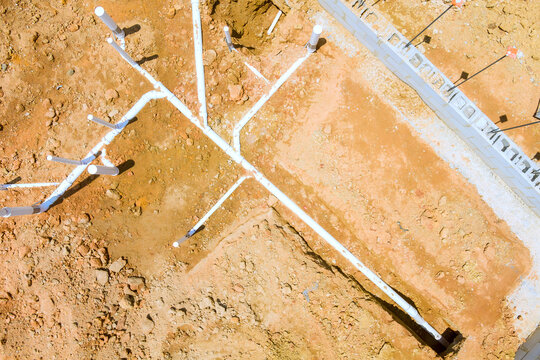 Workers are laying white pipes in fresh dirt at construction site under building foundation,