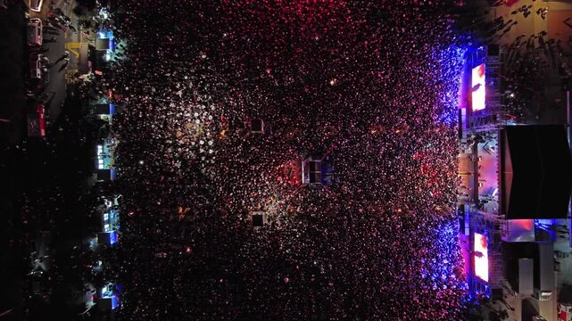 Aerial top view of crowd at night music concert. People gathered at festival outdoor stage with lights and screens, 4k