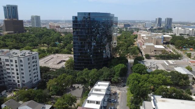 Tinted glass skyscraper overlooks dense tree canopies near Reverchon Park, Dallas. Surrounding parking garages complement expansive civic facilities nearby, DFW Metropolitan Area, Texas