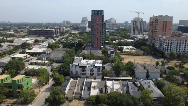 Fresh timber building framing rises beside residential townhomes near Oak Lawn, Dallas. Tall brick apartment highrises dominate sunny background skyline, DFW Metropolitan Area, Texas