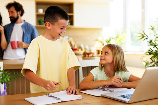 Brother and sister collaborate on homework at a kitchen table with notebooks and a laptop