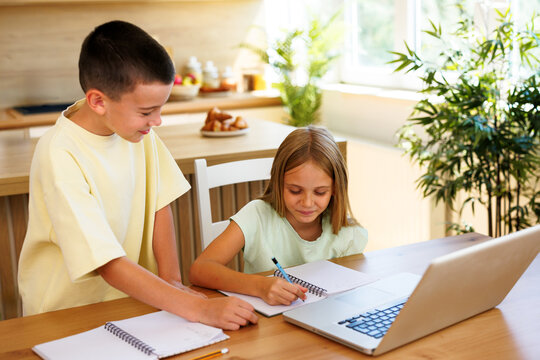 Two children work on school assignments at a kitchen table while their parents enjoy coffee together in the background