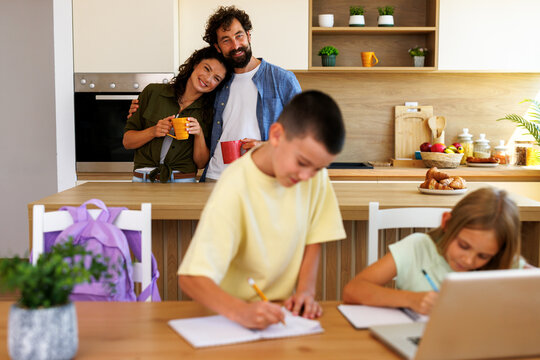 Two children study together at a kitchen table while their parents spend time together in the background