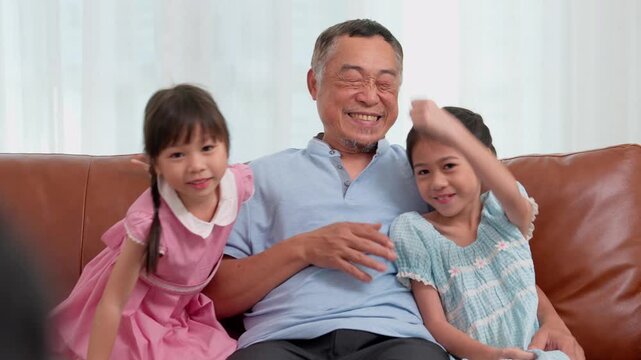 Grandfather spending playful time with two young granddaughters on a sofa at home. The family scene conveys affection, intergenerational bonding, happiness, and everyday family life.