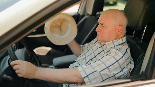 Elderly driver feeling hot and fanning himself with a hat inside the car.