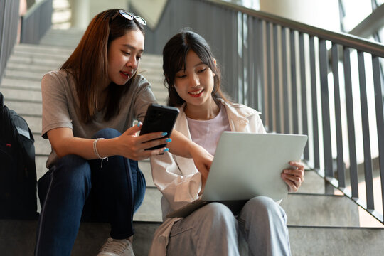 Two Asian Women sit on a staircase, reviewing smartphone and tablet together. The photo conveys collaboration, learning, and focus, highlighting teamwork, communication, and modern lifestyle