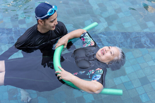 A male caregiver assists a senior woman floating with a pool noodle during aquatic rehabilitation in an outdoor swimming pool. The supportive interaction highlights recovery, mobility