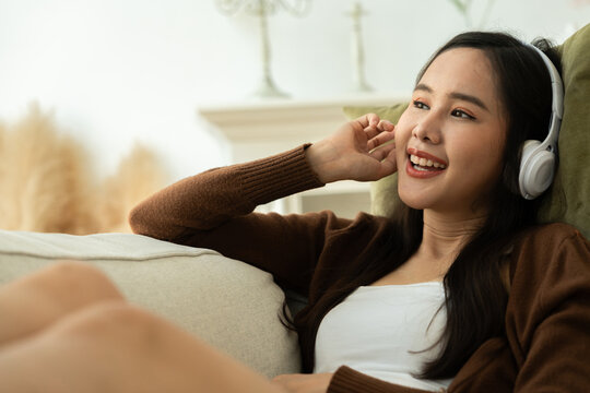 Relaxed and Joyful: Asian young woman in a cozy living room, headphones on, listening to music and enjoying the moment. Her relaxed posture and infectious smile convey happiness and contentment.