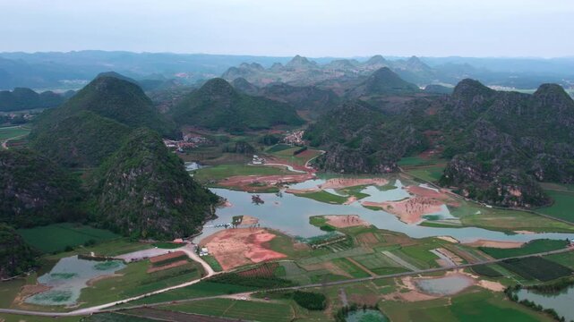Aerial view of a karst landscape with scattered water bodies, farmlands, and forested mountains under a pale sky.