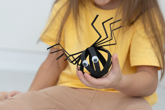Young Girl In Yellow Shirt Holds Handmade Black Paper Spider With Googly Eyes And Long Legs, Shown In Close Up Against Soft Neutral Background, Suggesting Playful Halloween Crafting