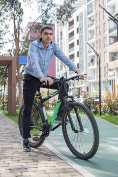 A young man is standing next to his bike on a pathway in a city. He wears a light blue shirt and black pants.