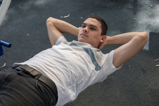 A young man does a workout routine using exercise equipment at an outdoor gym in a park during daylight.