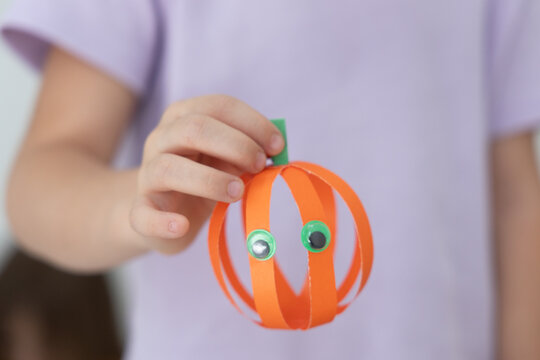 Close Up Of Small Hand Displaying Handmade Paper Pumpkin With Curved Orange Strips, Green Stem, Googly Eyes, And Soft Blurred Pastel Background, Suggesting Autumn School Craft, With Child, Hand