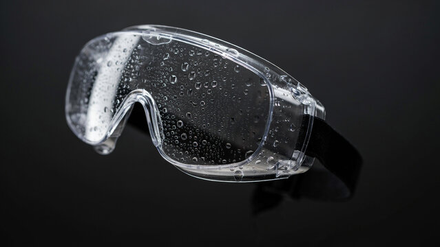 Clear protective safety goggles covered in water droplets with a black elastic strap isolated against a solid dark black background