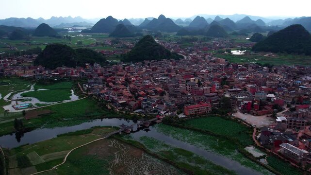 An aerial panorama shows a compact town nestled among karst peaks, with winding waterways and farmlands surrounding it.