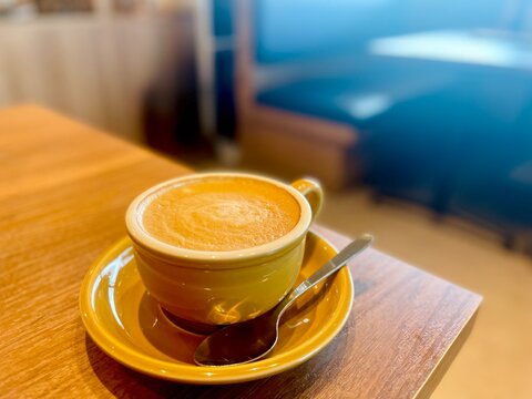 Warm creamy hot cappuccino in a yellow ceramic cup and saucer with a silver spoon on a wooden coffee shop table