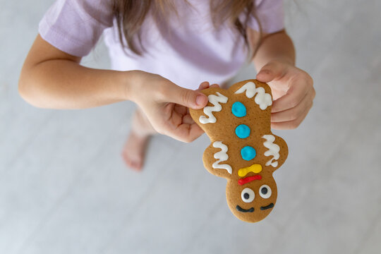 Young Child Holds Decorated Gingerbread Cookie Upside Down, Showing Colorful Icing Details, Pastel Shirt, Blurred Indoor Background, And Bright Natural Light For Festive Baking