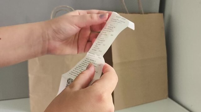 Woman checking a grocery receipt with paper shopping bags in the background, food expenses, rising prices, high grocery costs and household budget concept.