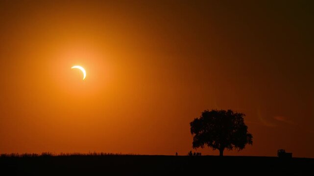 Time lapse with a spectacular view of a partial solar eclipse sunset with the silhouettes of a tree and people on the horizon watching the sun crescent in the deep orange sky. 