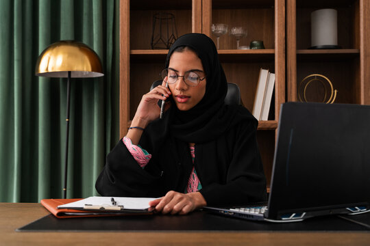 Middle-eastern businesswoman wearing abaya working in the office - focused arab businesswoman managing business in modern office while talking on phone