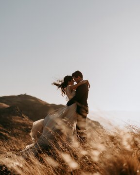 Bride and groom embracing on sunlit hilltop during golden hour with flowing dress and windblown grass, with bride, groom, embrace, wedding detail, showing couple, romance, love, outdoors context