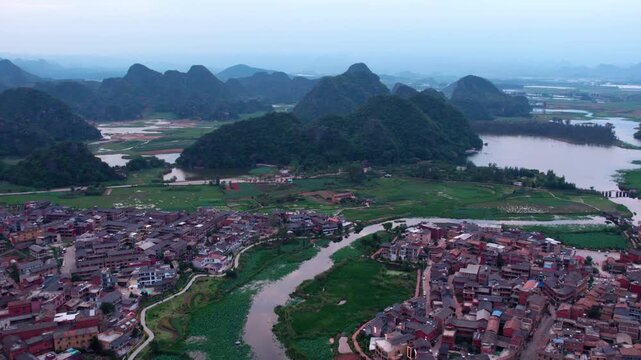 An aerial panoramic view of a riverside village nestled among karst mountains and lush green agricultural fields.