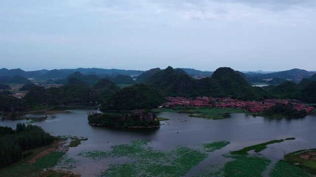 A panoramic landscape of a waterside village nestled among karst mountains, with aquatic plants covering the water surface under an overcast sky.
