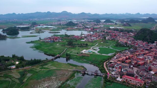 This aerial shot captures a water town with red roofs, surrounded by wetlands, fields and distant karst mountains under a pale sky.