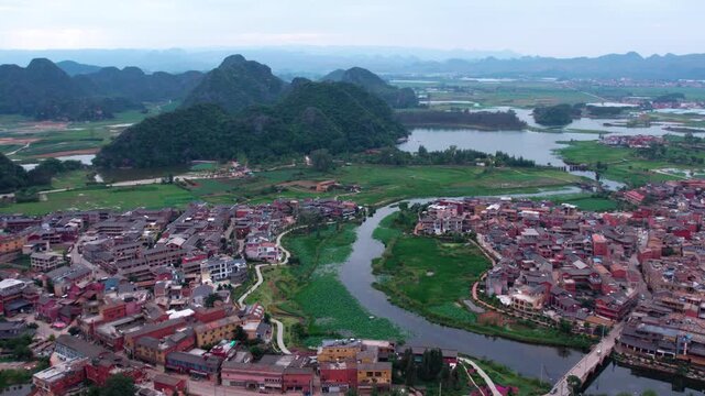 An aerial view of a riverside settlement surrounded by green karst mountains and wetland landscapes.