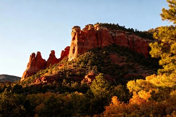 Naklejka premium Red rock formations rise above a forested landscape during autumn, illuminated by warm sunlight.