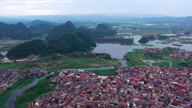 An aerial landscape showing a lakeside town nestled among forested karst mountains under a pale sky.