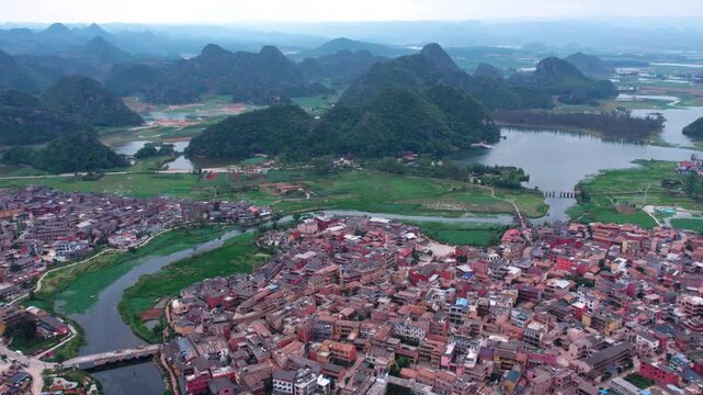 Aerial panorama of a riverside settlement nestled among karst mountains and water bodies, with dense buildings and natural landscapes.